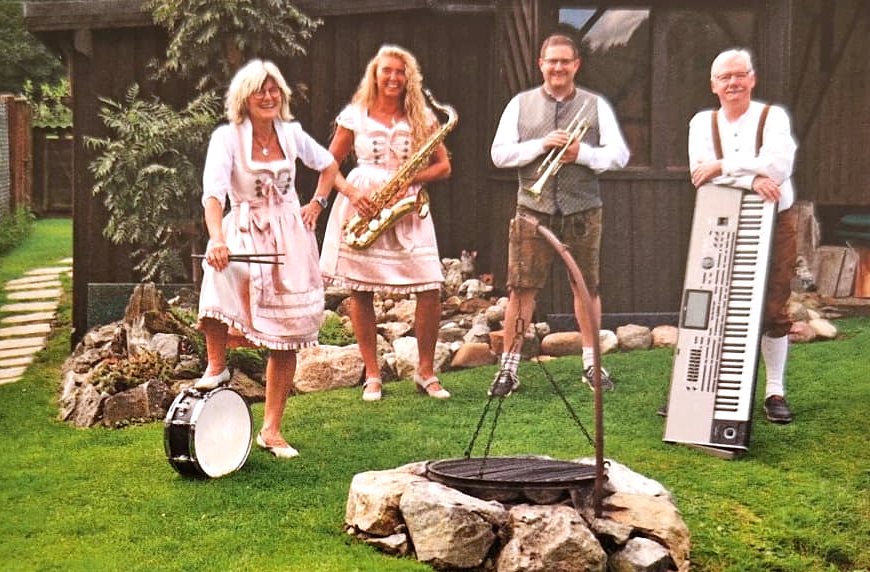 Die vier Musiker von a-round-music in Tracht im Garten: Schlagzeugerin mit Trommel, Saxophonistin, Trompeter und Keyboarder.